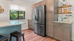 A modern kitchen with a stainless steel refrigerator, wooden cabinets, a marble countertop, and a window with a view of greenery.