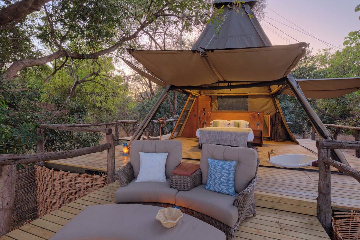 A tented accommodation with a wooden deck, cushioned seating, and a hot tub.