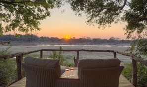 Una terraza de madera con mobiliario de mimbre y dos copas de vino, con vistas a un río y al atardecer.