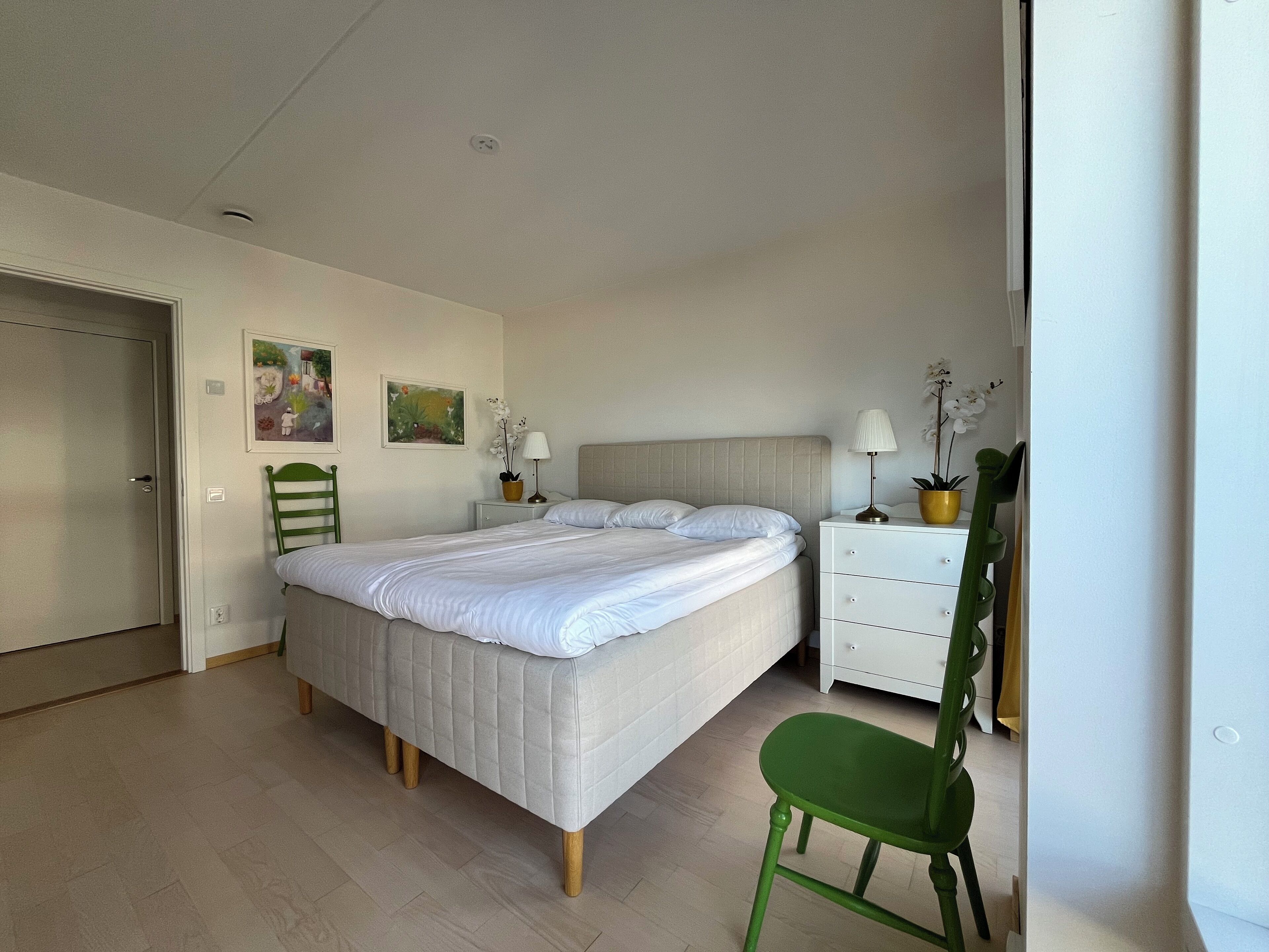 A bedroom with a bed, a green chair, a nightstand, a dresser, and two wall-mounted lamps.