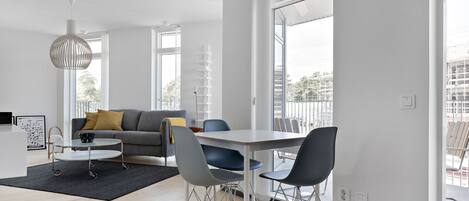 A modern living room with a grey sofa, a dining table with chairs, and large windows offering a view of the city.