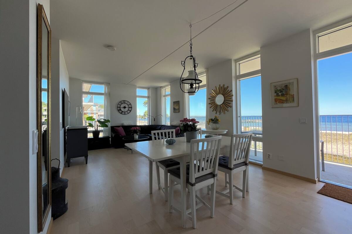 Een ruime woonkamer met een eettafel, witte stoelen, een zwarte bank en een groot raam met uitzicht op het strand.