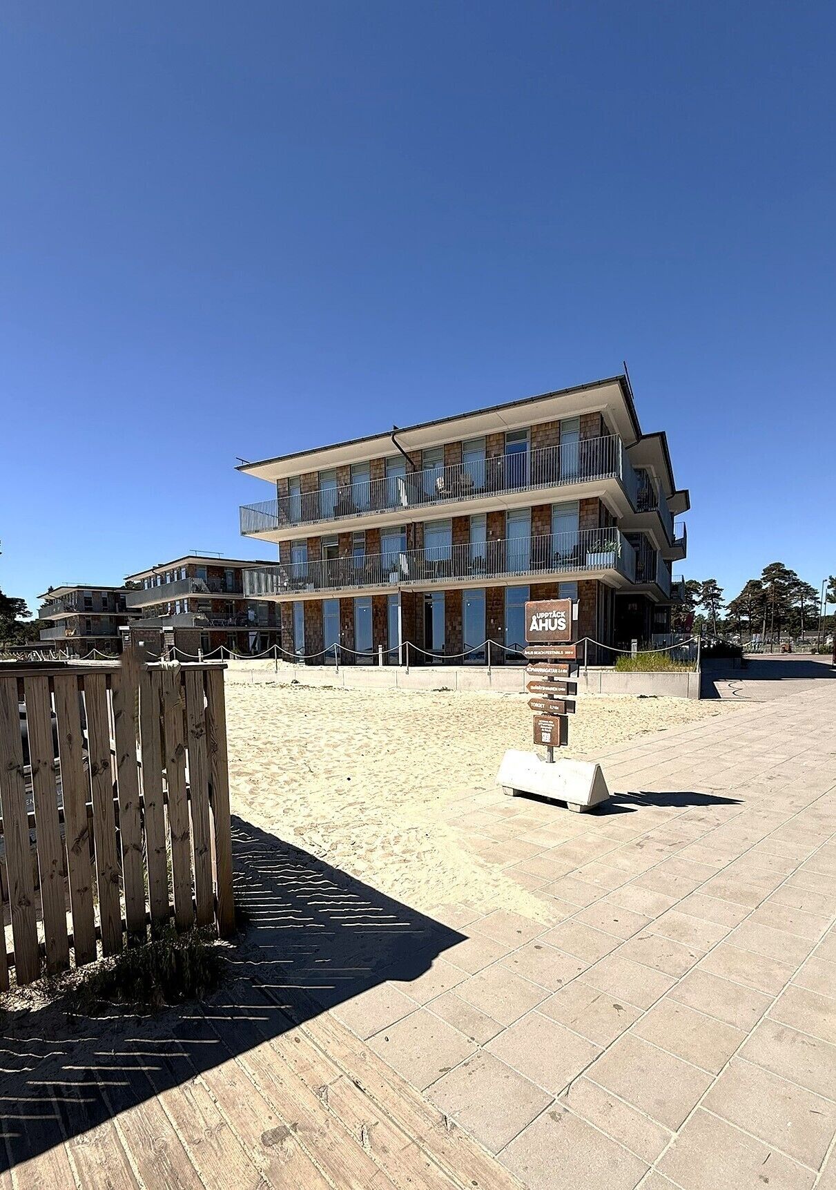Ein mehrstöckiges Hotel mit Balkonen, einer Sandstrandanlage und einem Holzzaun.