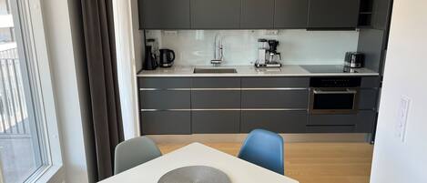 Una cocina moderna con armarios oscuros, encimera blanca y horno empotrado. Hay un comedor con mesa y sillas.