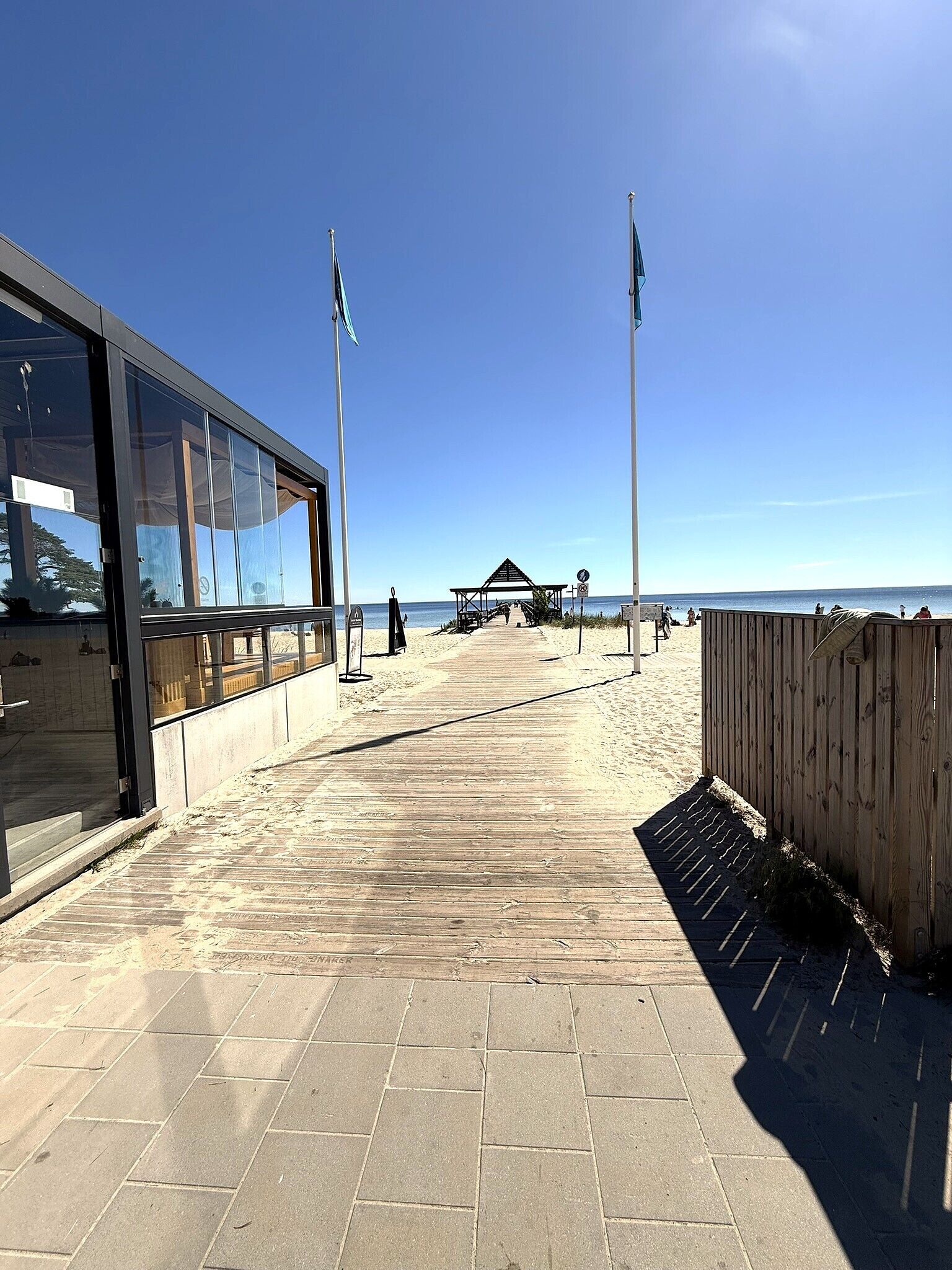 A beach walkway with a wooden fence, a covered seating area, and a building with large windows.