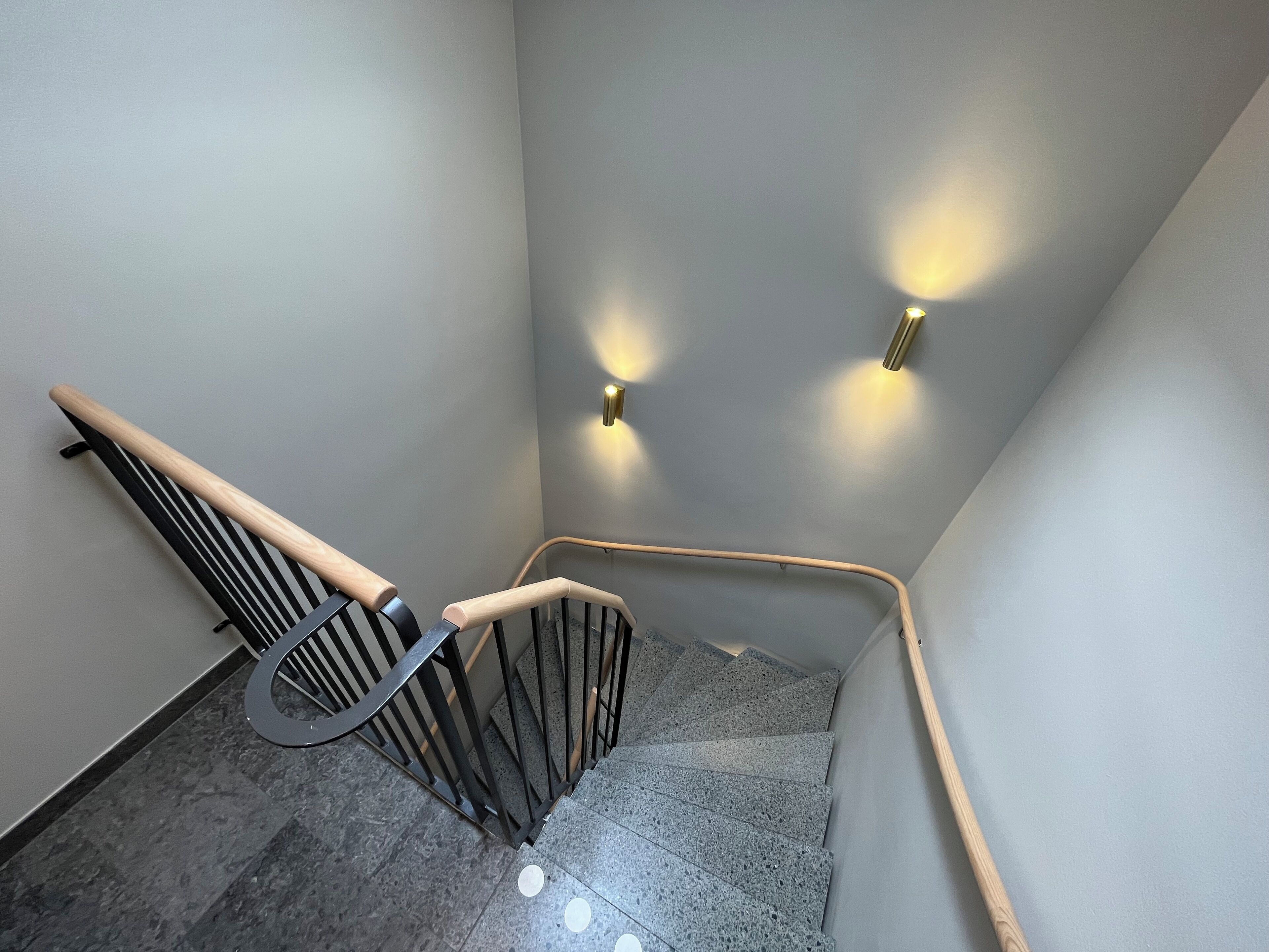 Un escalier moderne avec une main courante en bois et des balustrades en métal noir, menant à un palier agrémenté de deux appliques murales.