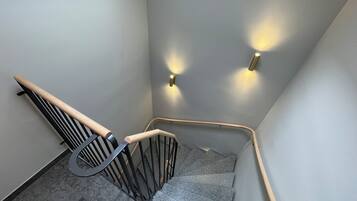 A modern staircase with a wooden handrail and black metal balusters, leading to a landing with two wall-mounted lights.