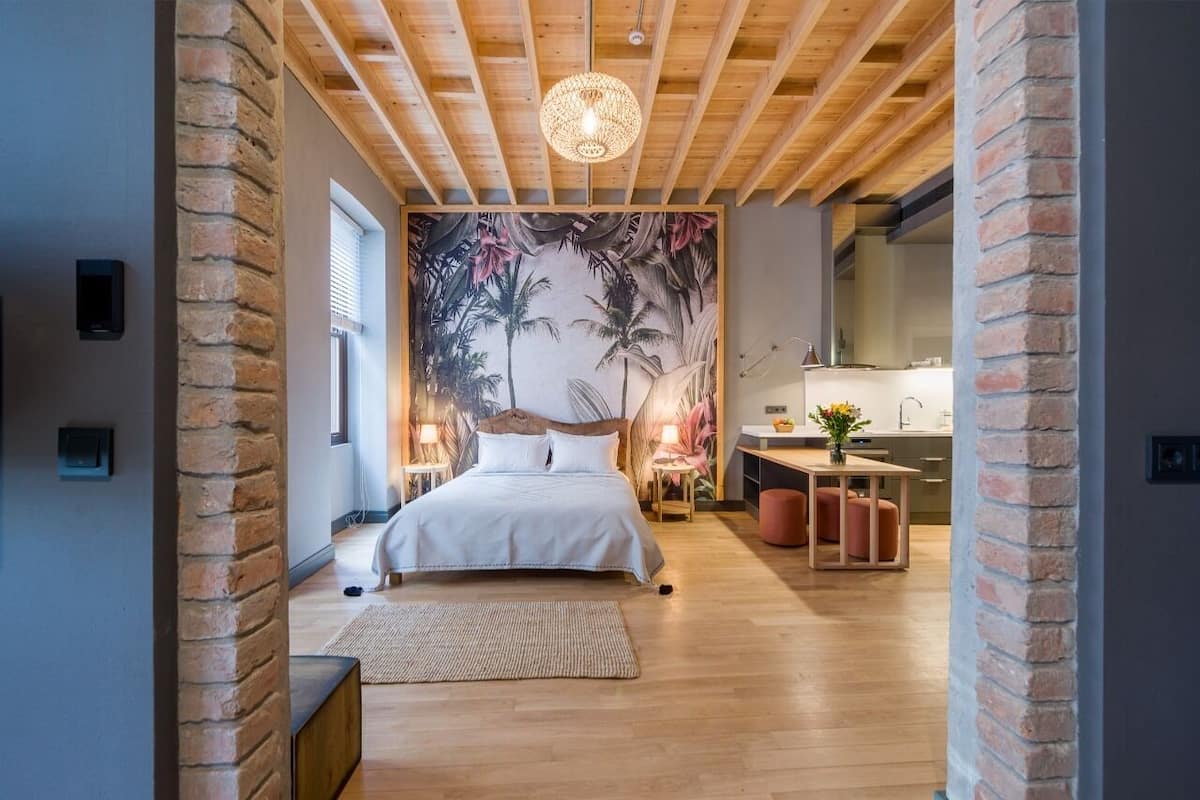 A bedroom with a bed, a large wall mural, a wooden ceiling, and a small table with a vase.