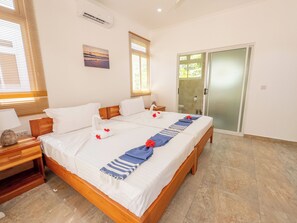 A hotel room with a large bed, a wooden headboard, a nightstand with a lamp, a painting on the wall, and a sliding glass door leading to a bathroom.