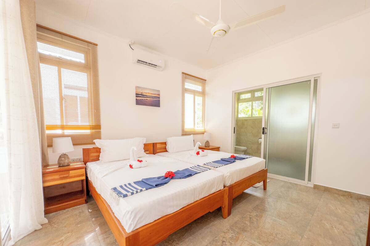 A double bed with a wooden frame, two bedside tables with lamps, a ceiling fan, and a sliding glass door leading to a bathroom.