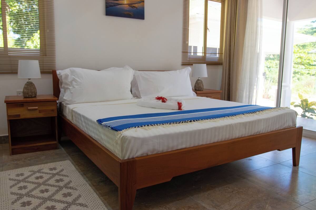 A neatly made bed with white linens and a blue and white striped blanket, a wooden nightstand with a lamp, and a patterned rug.