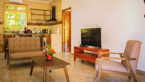 Una sala de estar con un sofá, una mesa de centro con flores, un televisor sobre un mueble bajo, y una cocina al fondo.