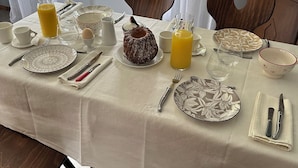 Una mesa de comedor dispuesta con platos, cubiertos, una jarra de jugo de naranja y un pastel, con vistas al follaje exterior.