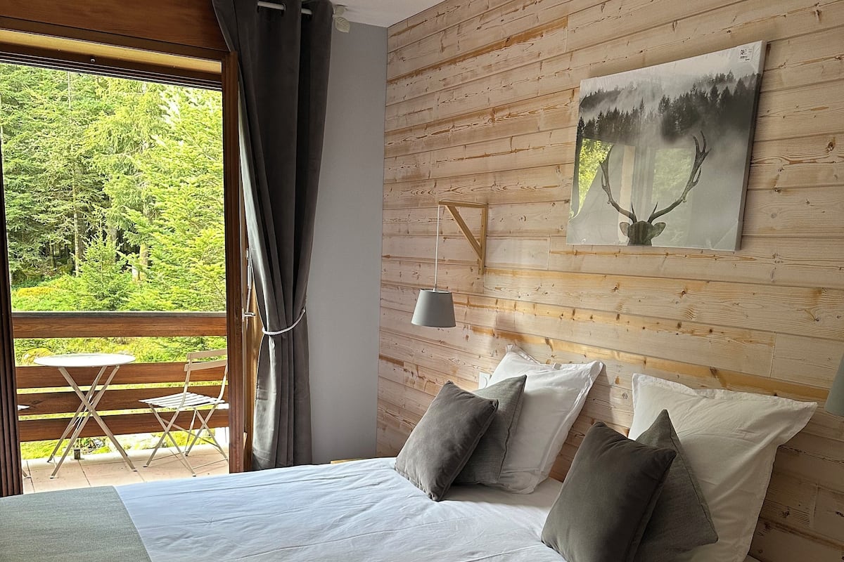 A bedroom with a large bed, wooden headboard, a painting of a deer, and a view of a forest through a window.