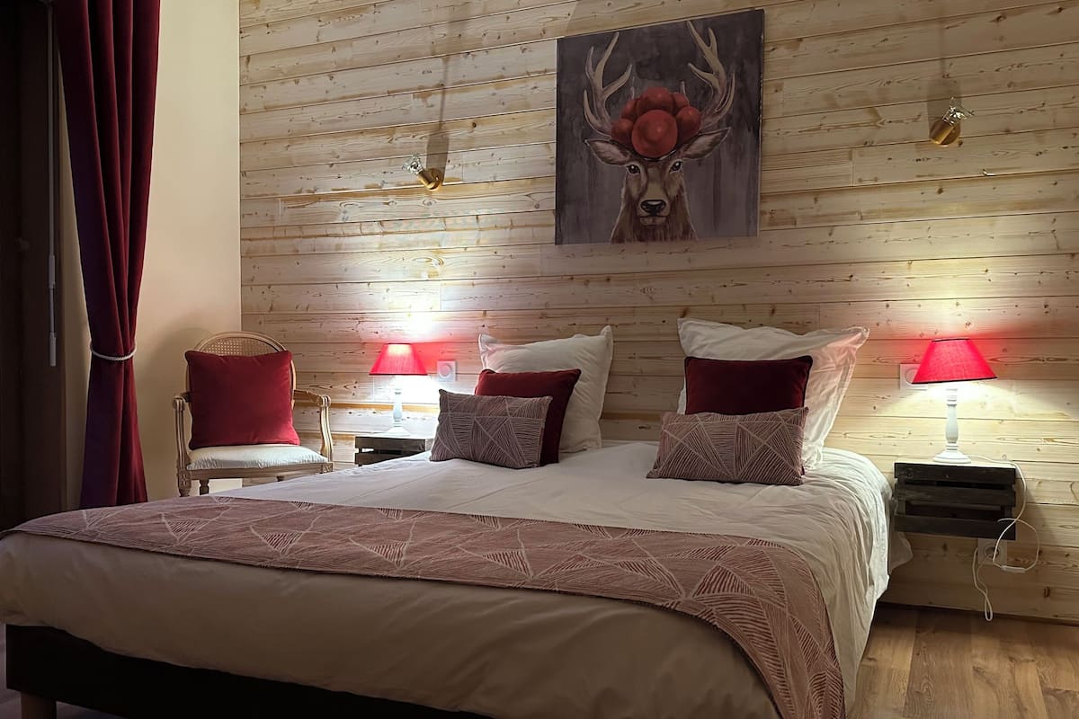 A bedroom with a wooden wall, a bed with multiple pillows, two bedside tables with lamps, and a painting of a deer.
