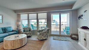 Ein geräumiges Wohnzimmer mit blauem Sofa, gemusterten Sesseln, einem runden Couchtisch und Blick auf den Balkon mit Gartenmöbeln.