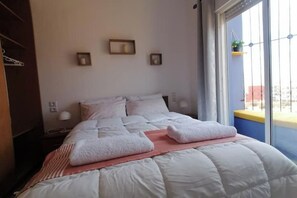 Una cama bien hecha con ropa de cama blanca y dos toallas dobladas, una mesita de noche con lámpara, y una ventana con vistas al exterior.