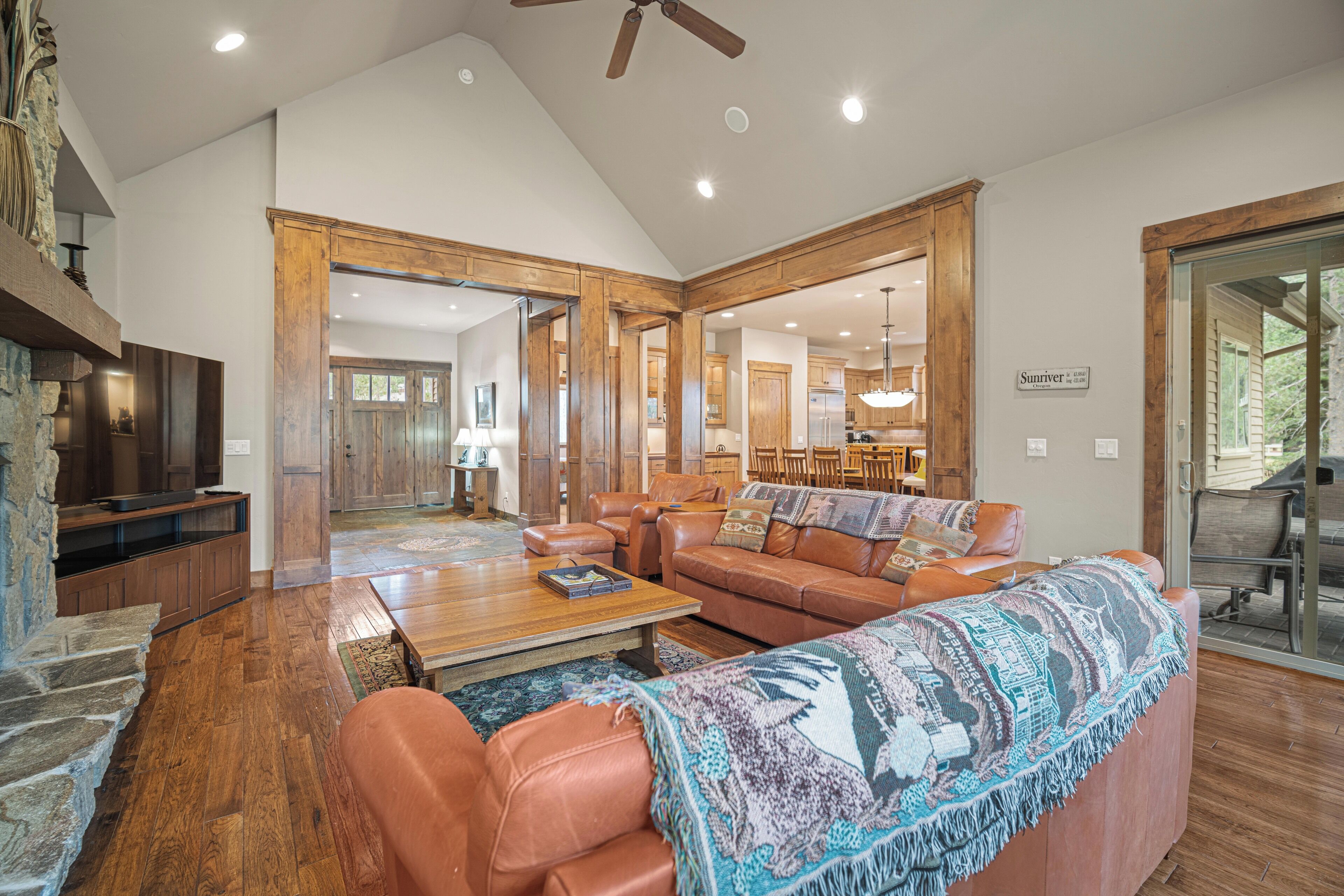 A spacious living room with a large sofa, a coffee table, and a television. There is a wooden ceiling fan and a stone fireplace.