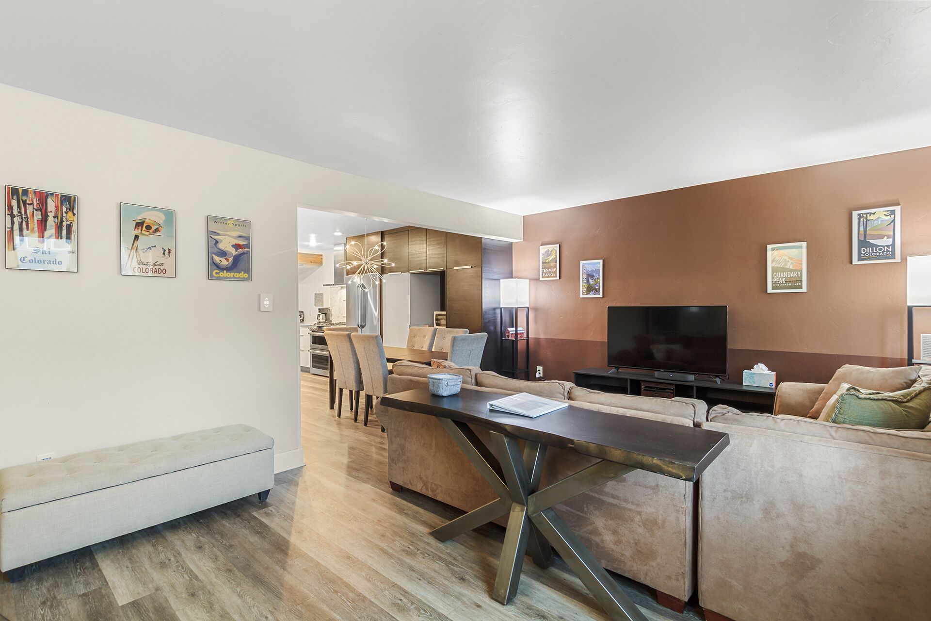 A modern living room with a sofa, a dining table, and a TV. There are posters on the walls and a bench in the corner.