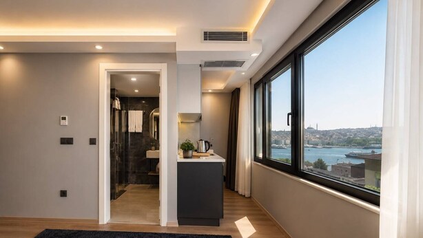 A modern interior with a view of a cityscape through large windows, a kitchen area, and a doorway leading to a bathroom. - ART Karakoy Hotel (Istanbul)