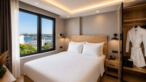 A modern hotel room with a large bed, bedside lamps, a wooden headboard, a view of the city, and a robe hanging on the door. - ART Karakoy Hotel (Istanbul)