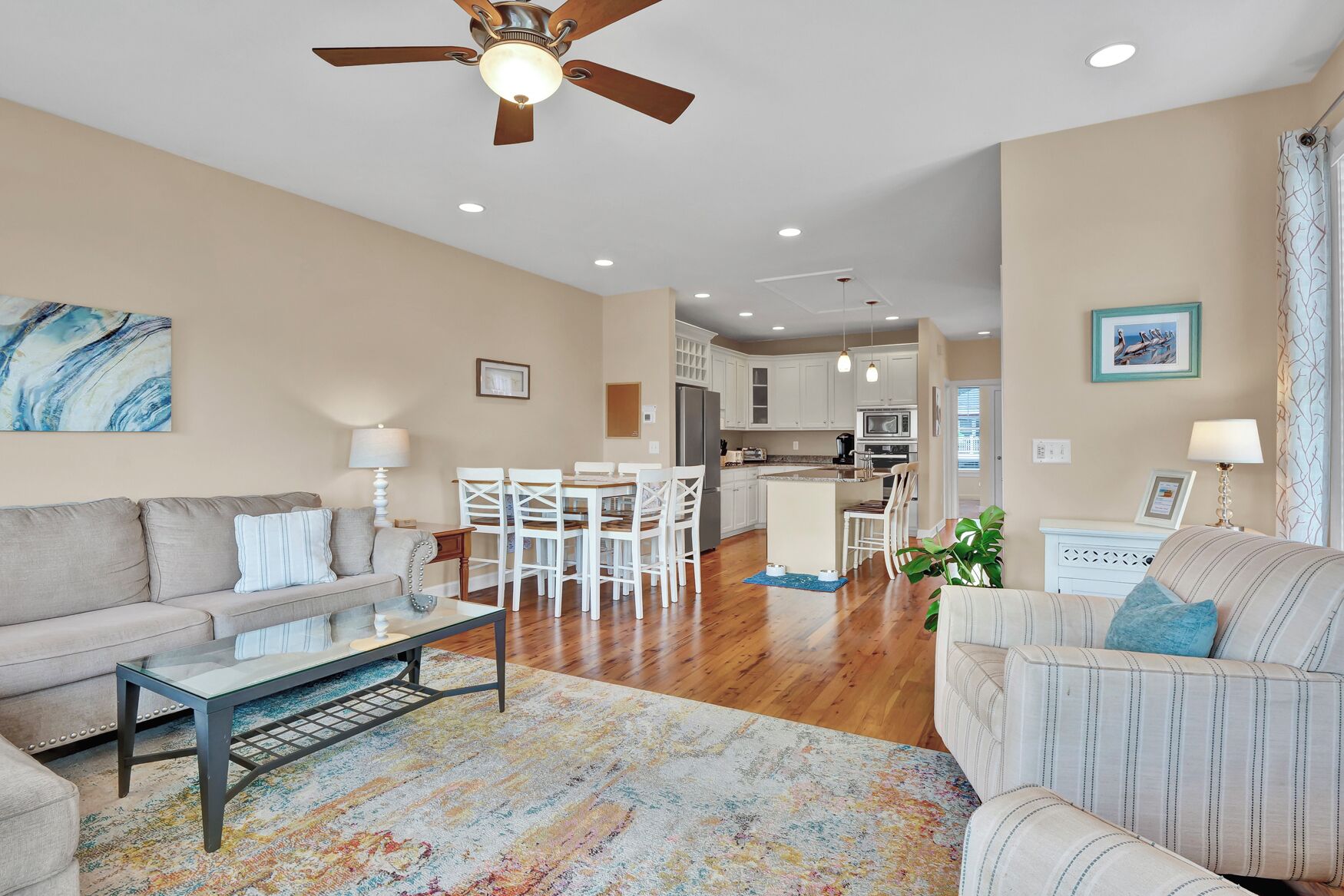 A spacious living room with a sofa, coffee table, and a dining area with chairs. A ceiling fan and recessed lighting are visible.