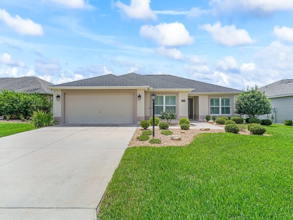 A single-story house with a garage, a well-maintained lawn, and a small garden with shrubs and a tree. - Stay in Style near the Golf Course in the Village of Fenney! (The Villages)
