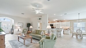 A spacious living room with a green sofa, a coffee table, and a dining area with white chairs. There is a kitchen in the background with white cabinets and a ceiling fan. - 3/2 Designer with a Golf Cart, close to Lake Sumter & Brownwood!!! (The Villages)