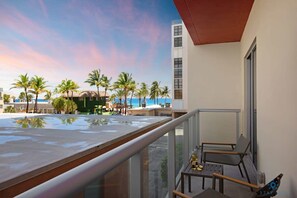 Un balcón con vista a una piscina, palmeras y un edificio con techo rojo.