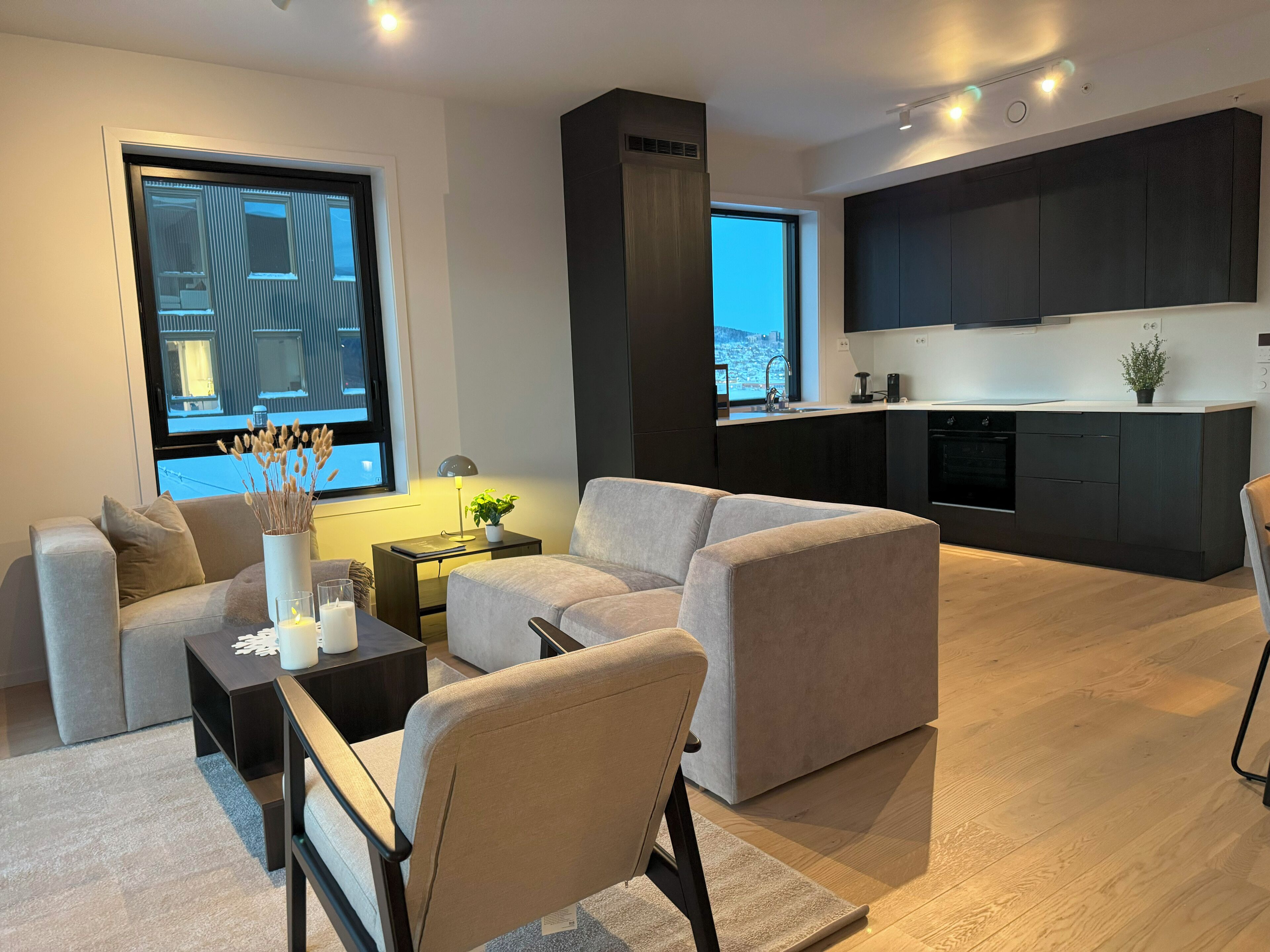 Un salon moderne avec un canapé, une table basse et une petite table d’appoint sur laquelle se trouvent une lampe et une plante. Une grande fenêtre offre une vue sur un immeuble.