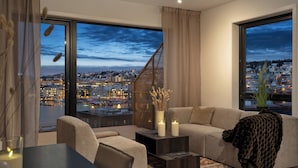 A modern living room with a large window, a sofa, a coffee table, and a view of a cityscape at dusk. - TotalApartments Vervet Gjøa (Tromsø)