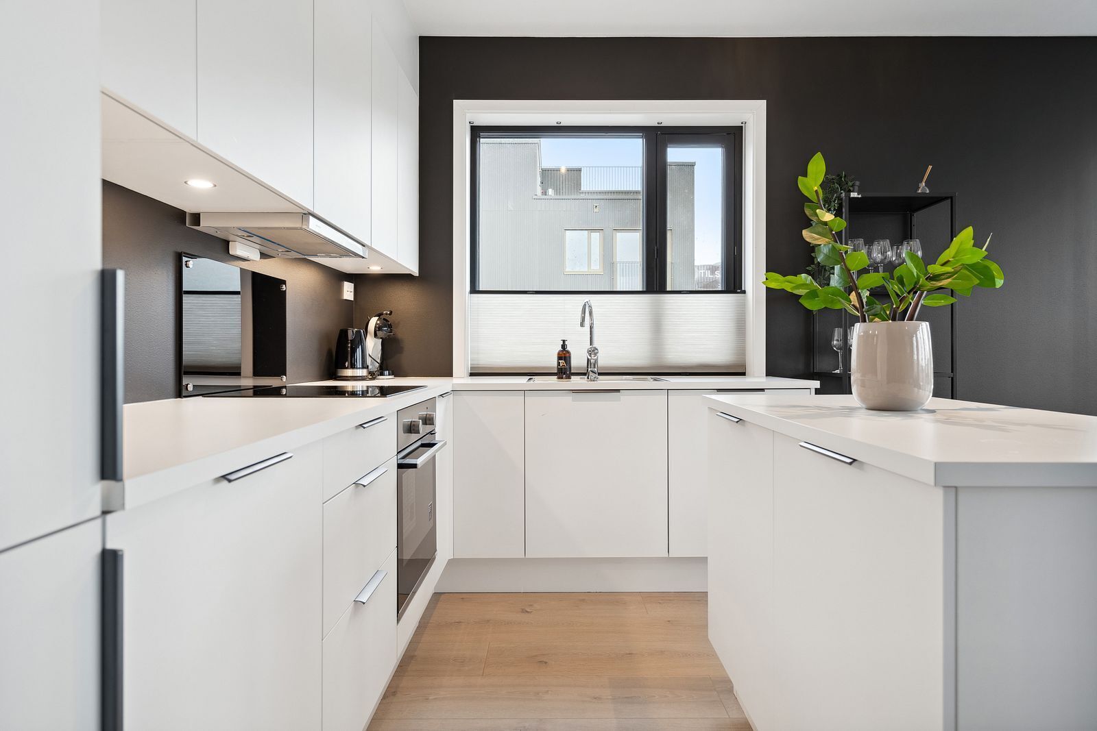 A modern kitchen with white cabinetry, a stainless steel range hood, a built-in oven, and a window above the sink.