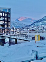 Un área ribereña cubierta de nieve con un puente, edificios y montañas al fondo.