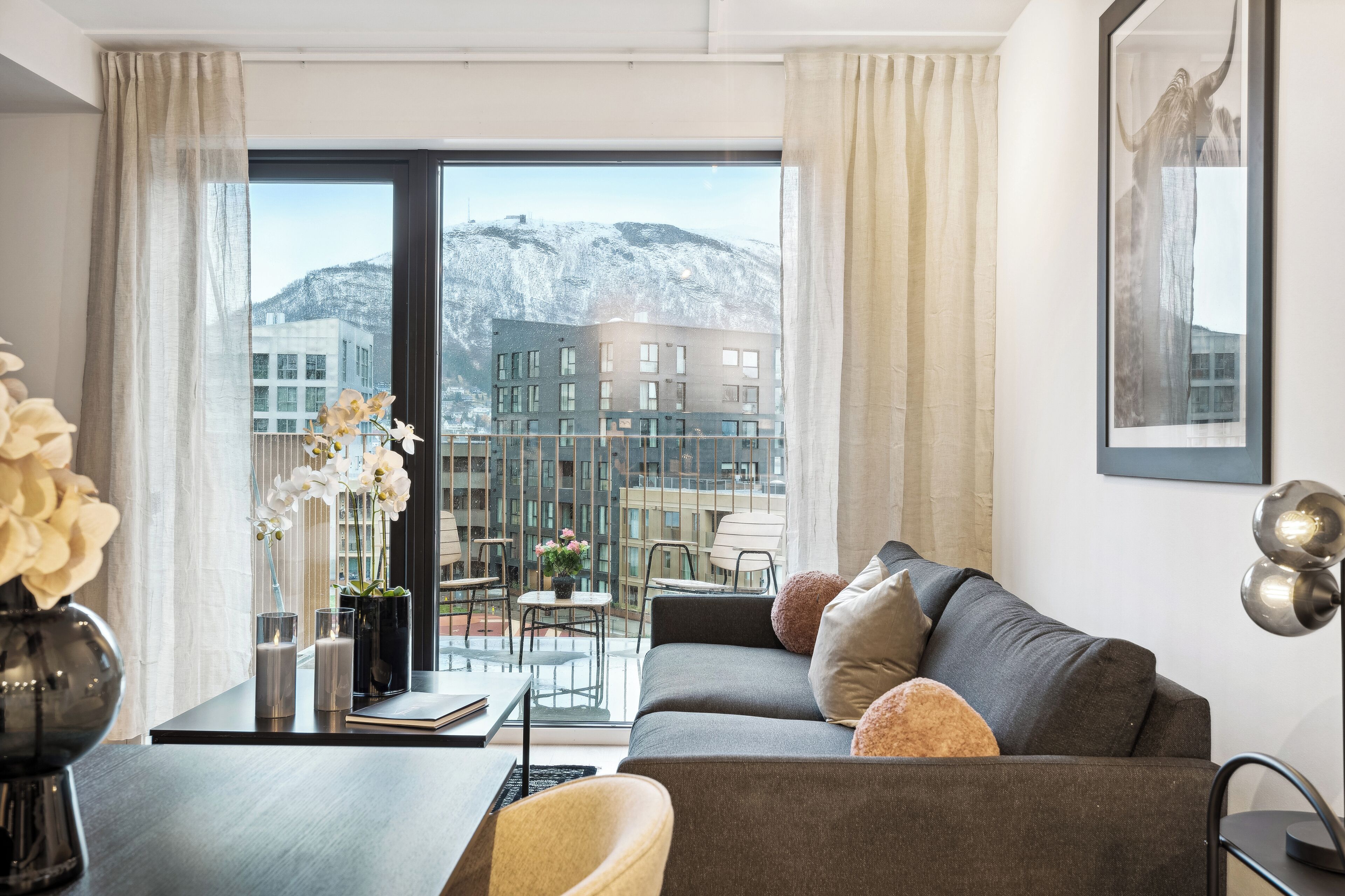 A modern living room with a grey sofa, a small table with a vase of flowers, and a view of a snowy mountain through large windows.