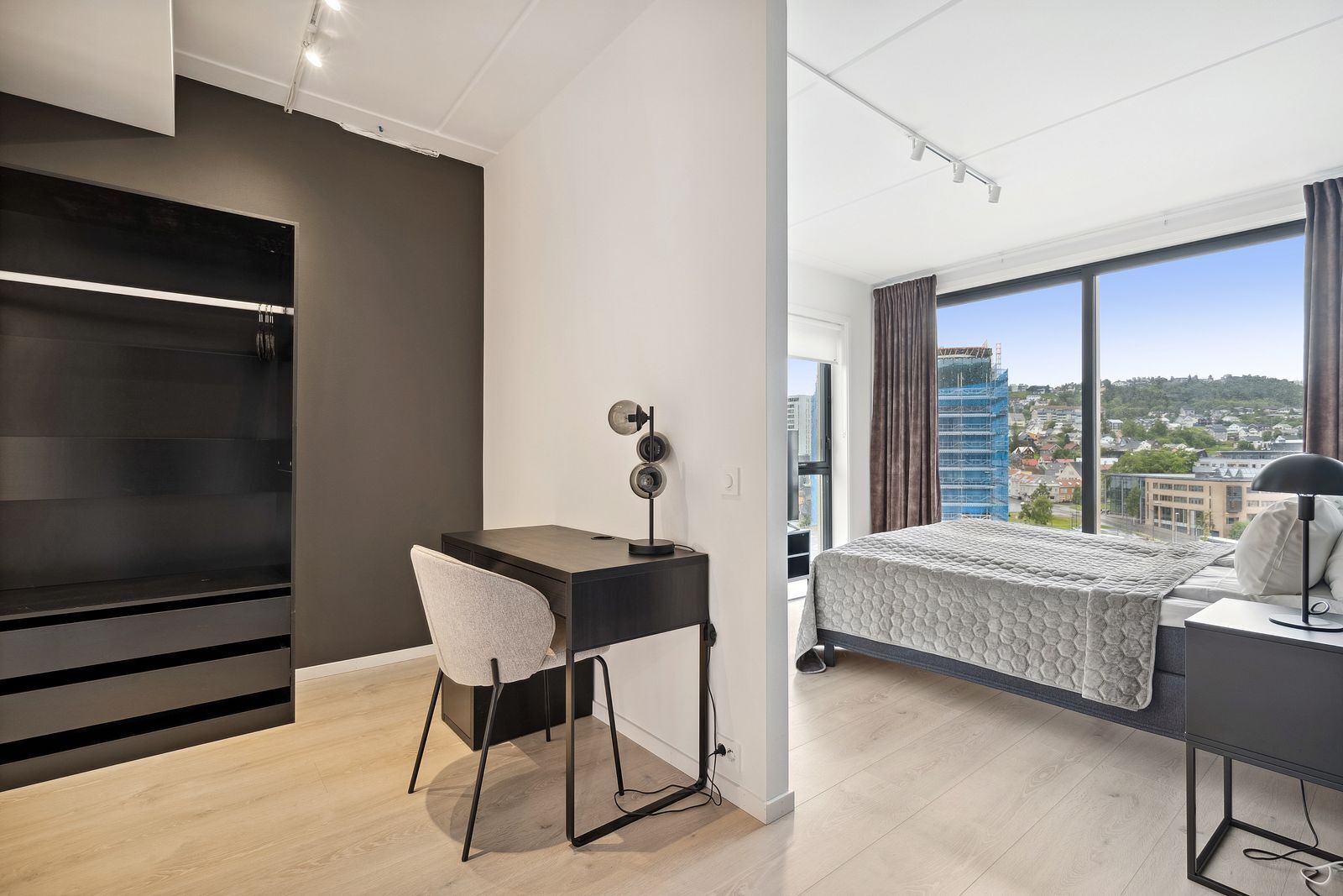 Une chambre moderne équipée d’un bureau, d’un lit et d’une grande fenêtre donnant sur la ville.