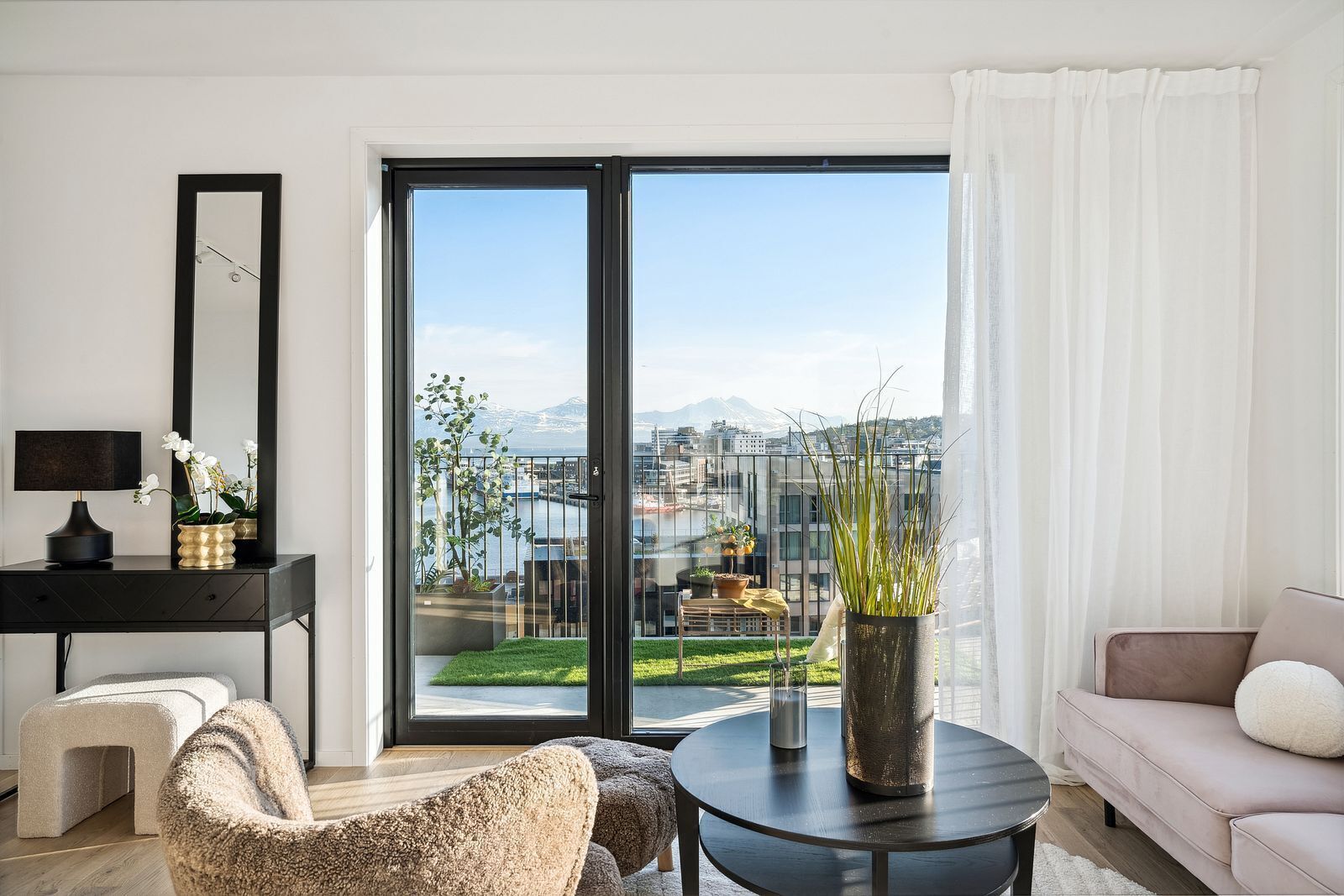 A modern living room with a large sliding glass door, a sofa, a coffee table, a desk with a lamp, and a view of a cityscape and mountains.