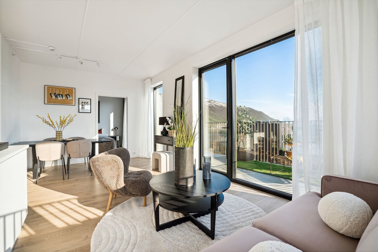 A modern living room with a large sliding glass door leading to a balcony, a dining area with a table and chairs, and a kitchen area.