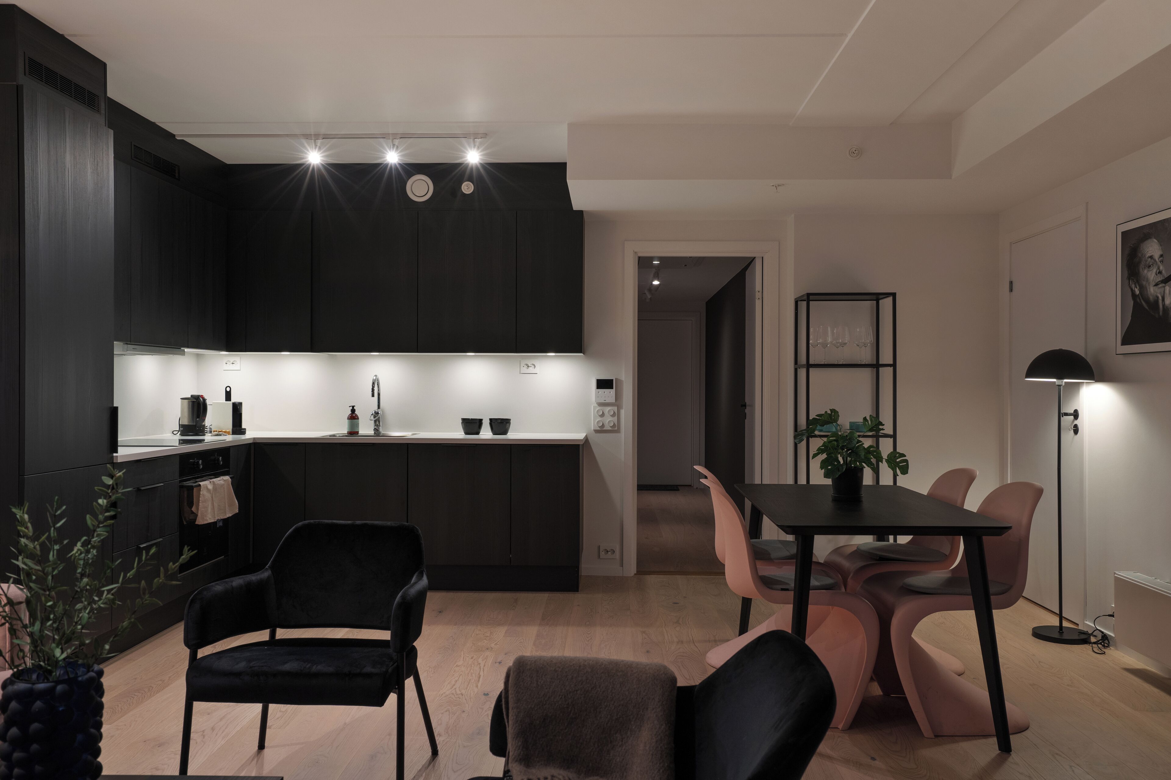 Una cocina moderna con armarios oscuros, una isla central e iluminación colgante. Hay una zona de comedor con una mesa y sillas, y una planta en maceta sobre la mesa.