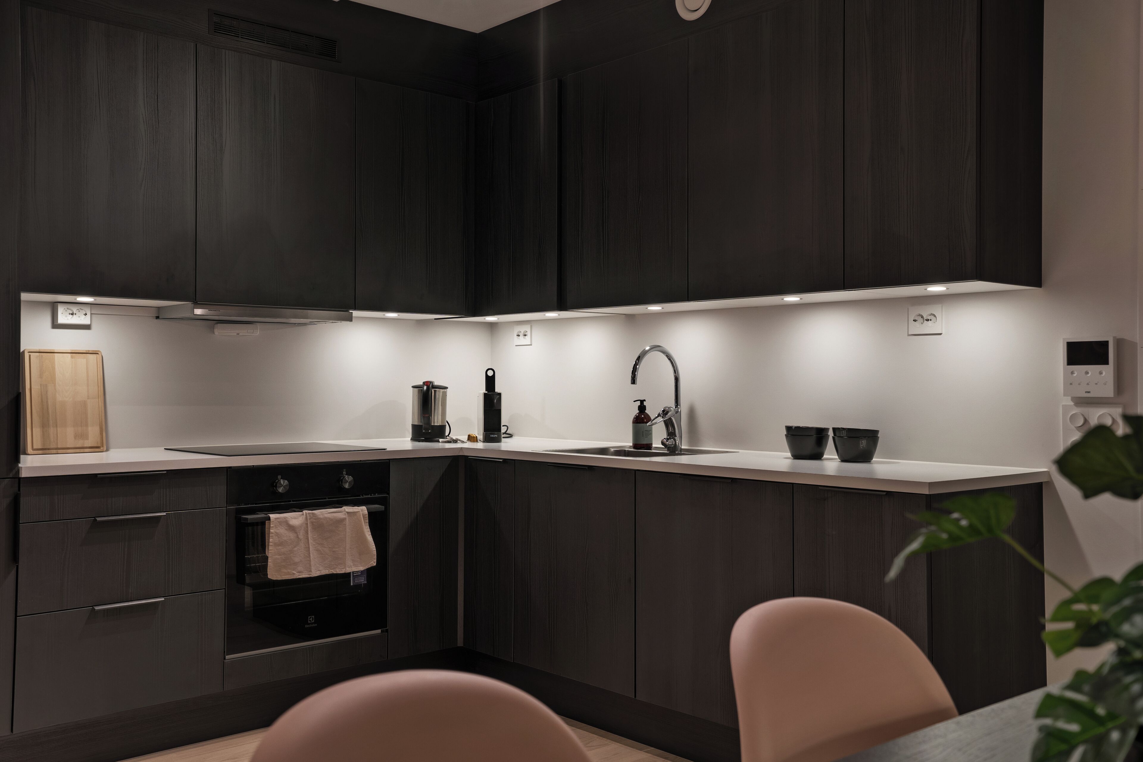 Une cuisine moderne avec des meubles de couleur bois foncé, une hotte en acier inoxydable, un évier double et un petit coin repas avec deux chaises.