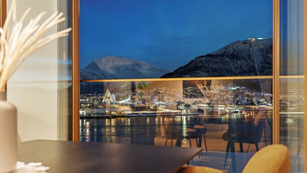 A view from a hotel room with a large window overlooking a cityscape at night, featuring a body of water, city lights, and a mountain in the background. - TotalApartments Vervet Gjøa (Tromsø)