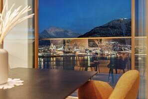 A view from a hotel room with a large window overlooking a cityscape at night, featuring a body of water, city lights, and a mountain in the background. - TotalApartments Vervet Gjøa (Tromsø)