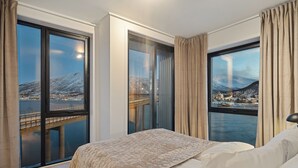A hotel room with a large bed, a view of a cityscape through a window, and a balcony with a view of a bridge and mountains. - TotalApartments Vervet Gjøa (Tromsø)