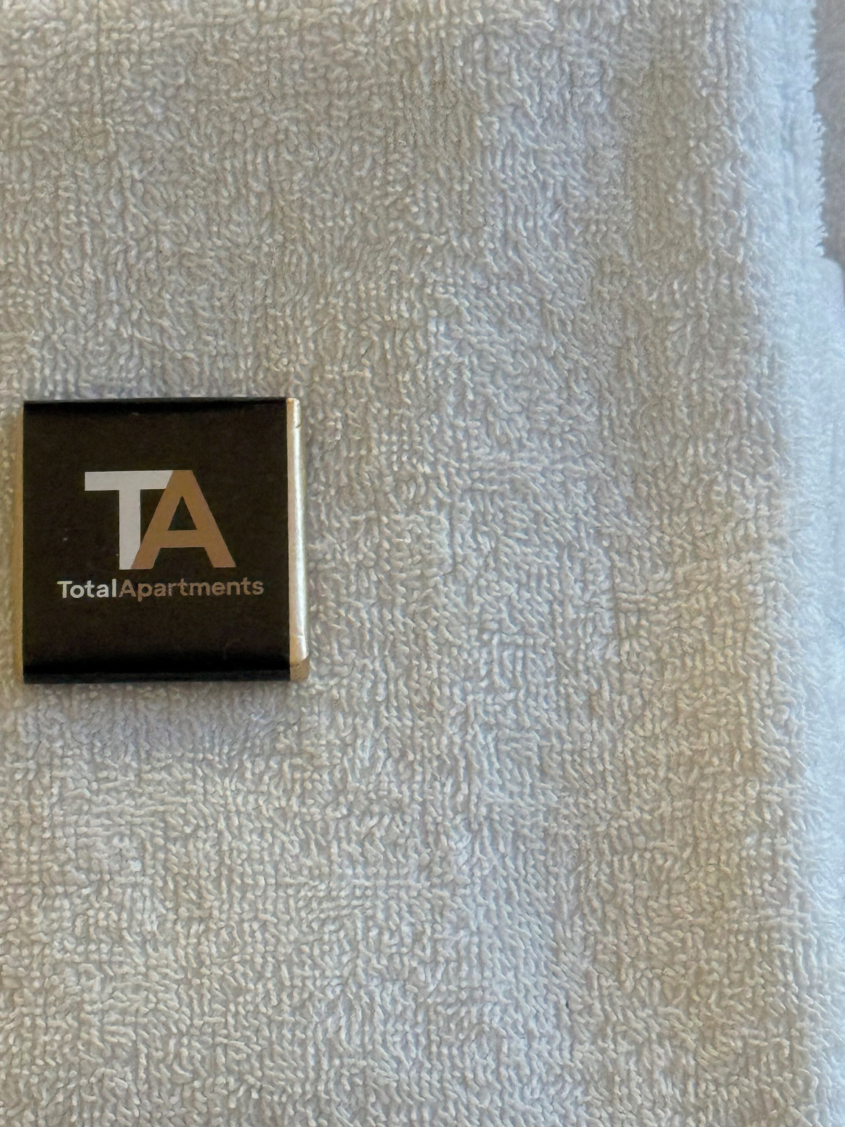 Een klein zwart bord met het logo en de naam 'TotalApartments' op een gestructureerd wit oppervlak.