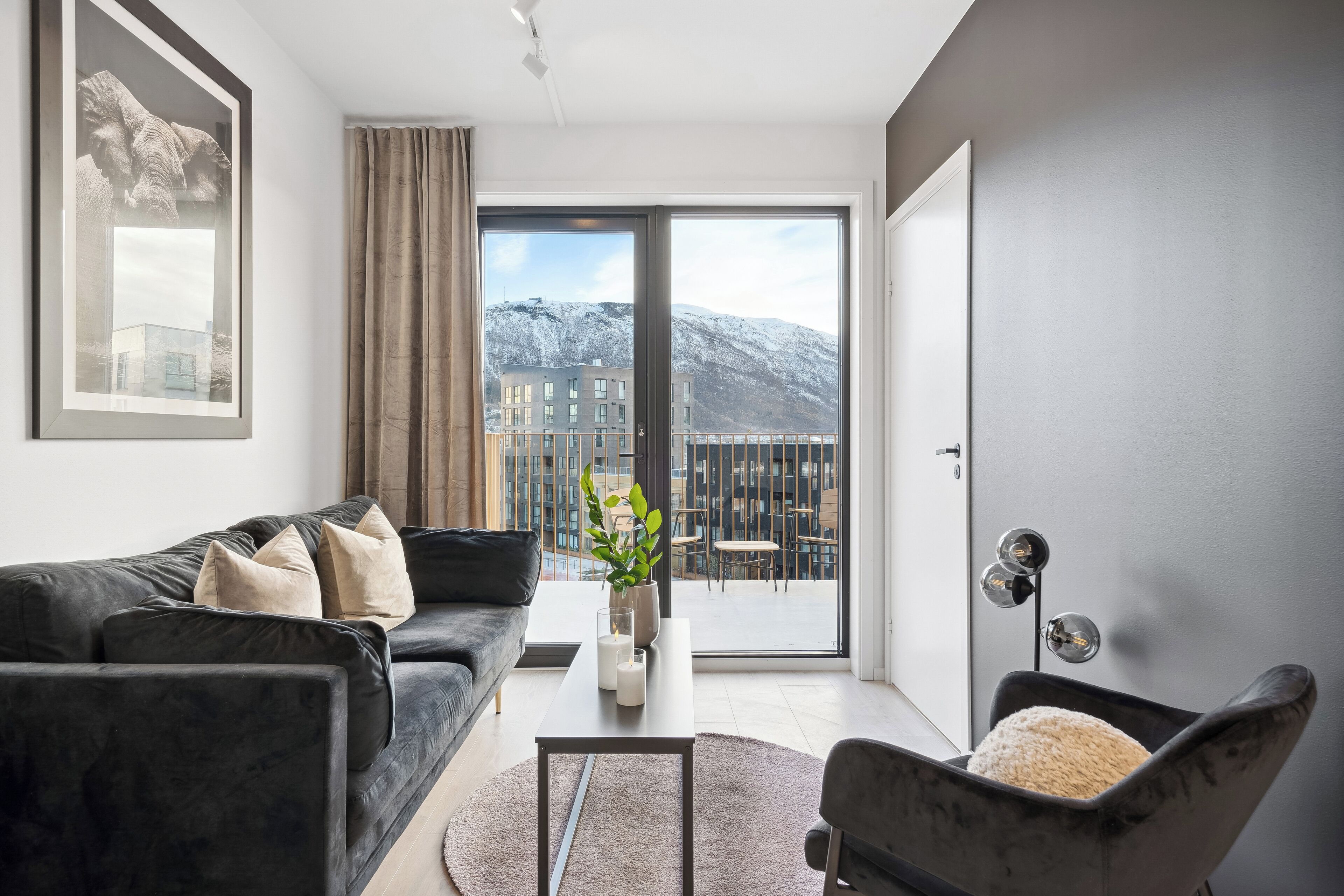 A modern living room with a black sofa, a small table with a vase and candles, a chair with a cushion, and a large sliding glass door leading to a balcony with a mountain view.