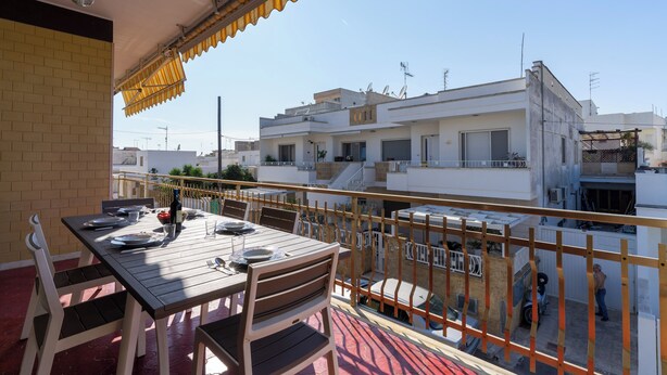 Varanda com mesa posta para refeições, com vista para área residencial com edifícios brancos e céu claro.