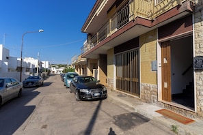 Vista da rua com carros estacionados, um edifício com varanda e céu azul claro.