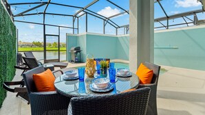 Um pátio externo com mesa de vidro posta para refeições, rodeado por cadeiras de vime e copos de vidro azuis.