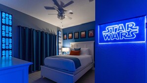 Quarto com cama, ventilador de teto, parede com tema Star Wars e mesa de cabeceira com abajur.