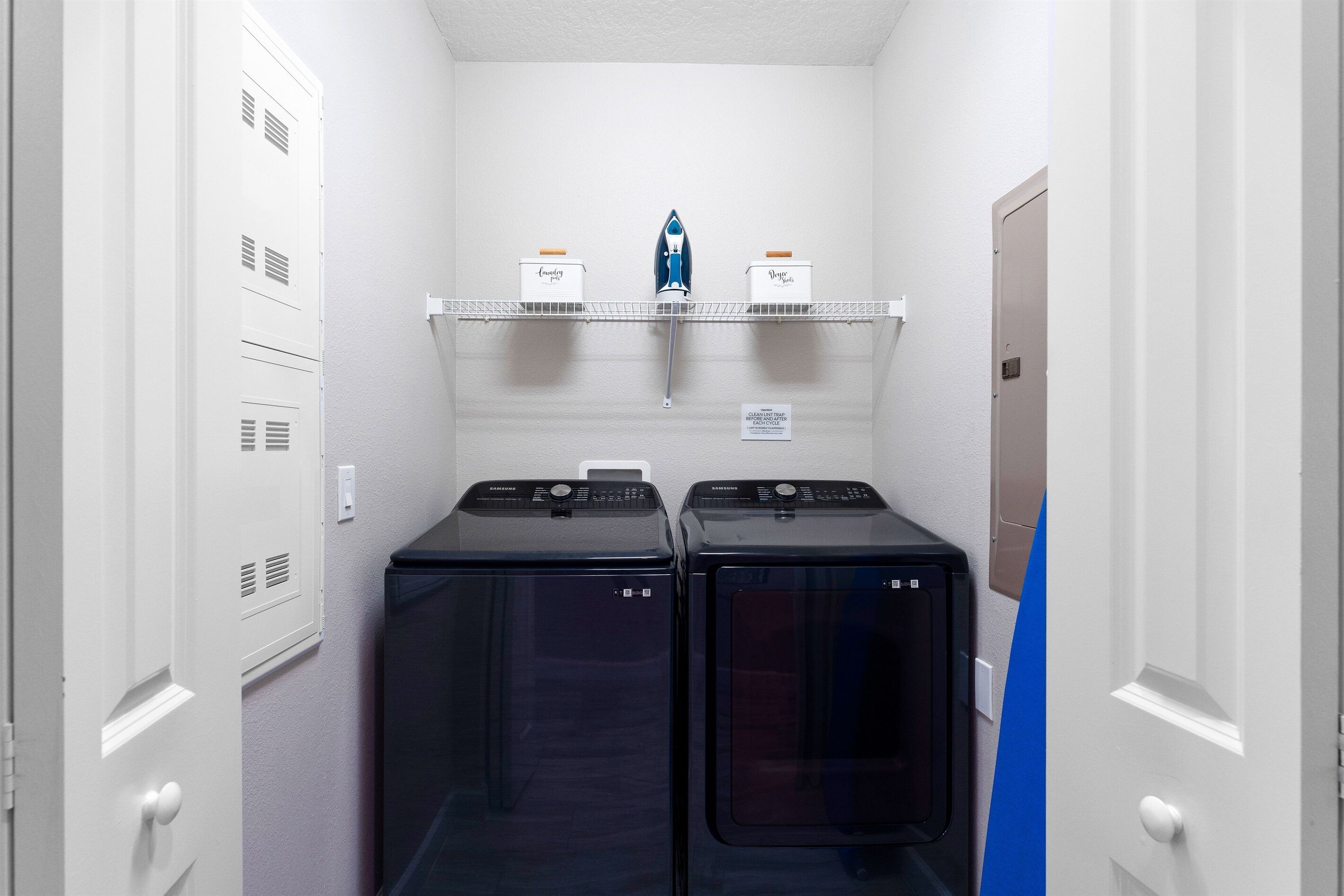 A laundry room with two front-loading washers and dryers, a shelf with ironing supplies, and a door to the right.
