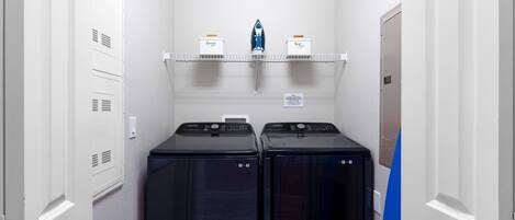 A laundry room with two front-loading washers and dryers, a shelf with ironing supplies, and a door to the right.